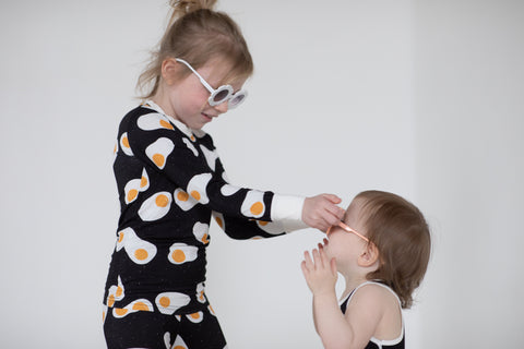 An older sister wearing egg pjs and sunglasses places sunglasses on her younger sisters face