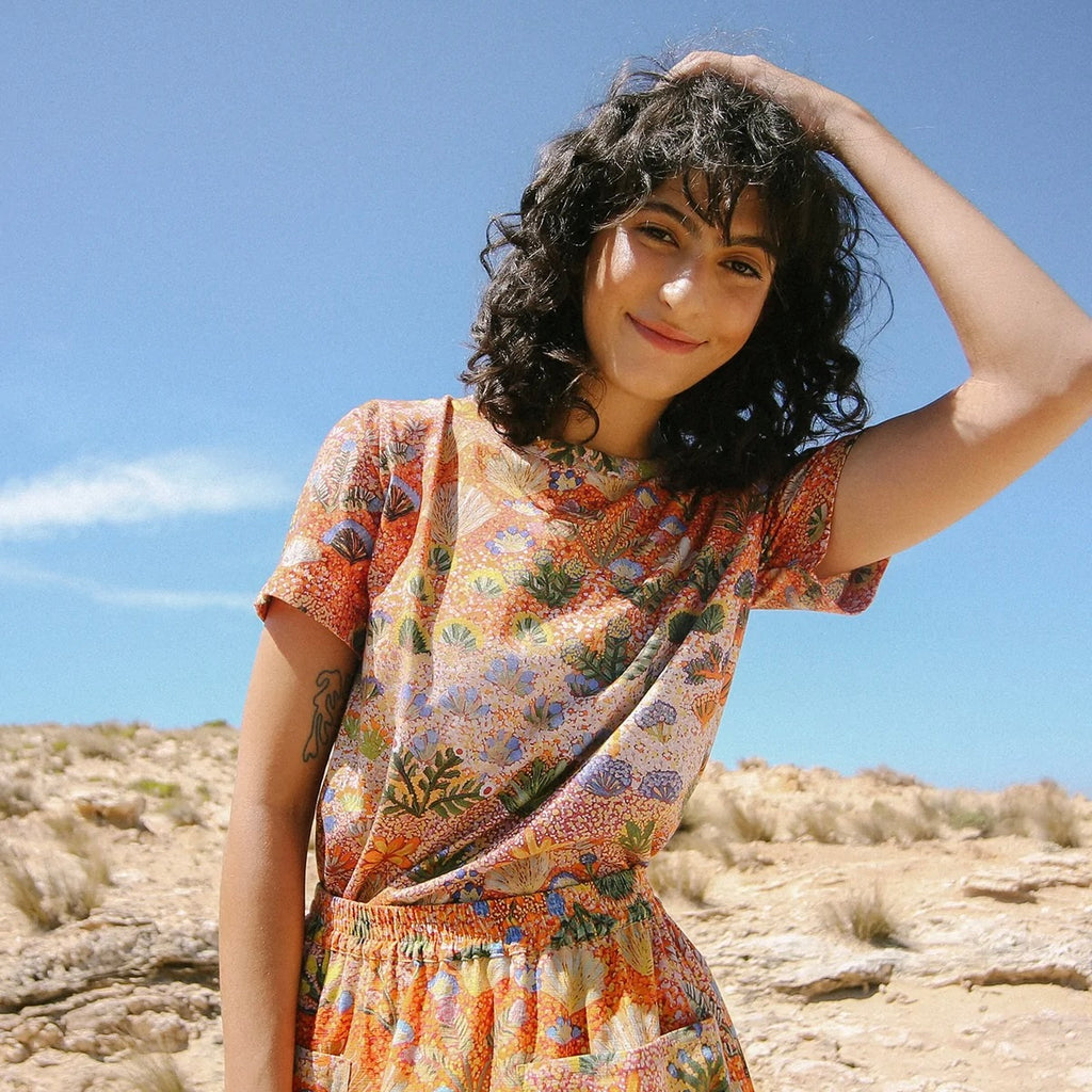 Mara wears the Nancybird Apollo Tee in Desert Flower print while smiling at the camera with her mouth closed. She has curly, shoulder-length dark hair. The background is a desert landscape with native grasses and a blue sky with a couple of wispy clouds.