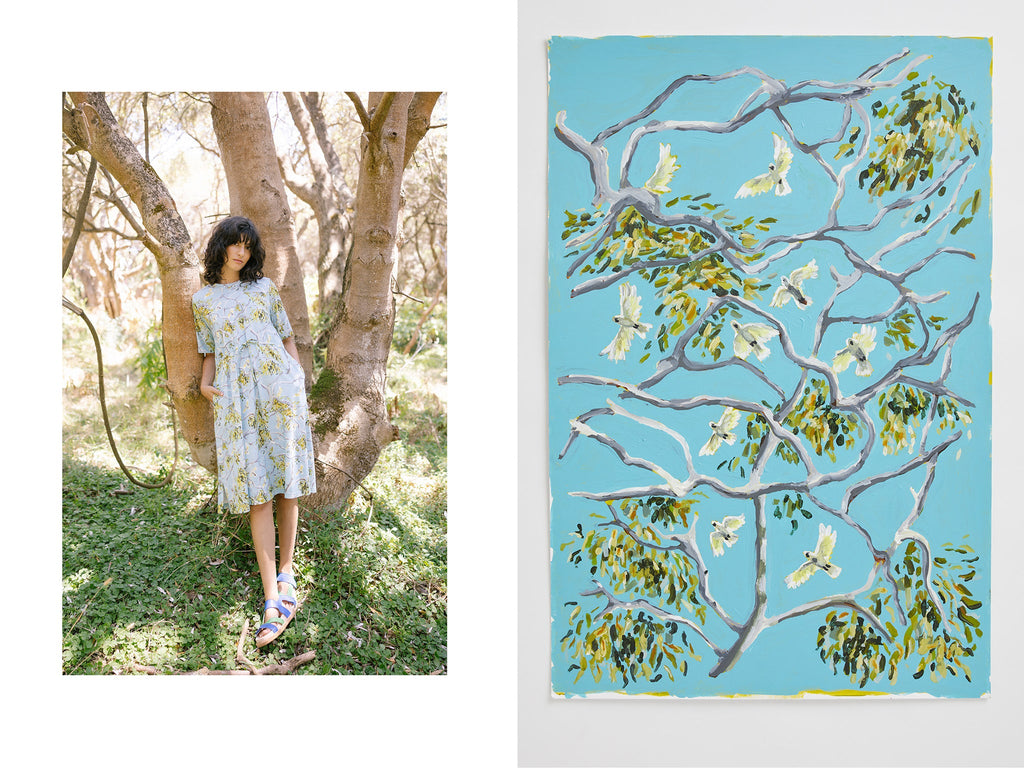 A woman with curly dark hair leans against a tree wearing the Nancybird Pepper Dress in Treetops which has a blue background with gum branches, leaves and cockatoos printed on it. To the right of the photo is the original artwork that the print of the dress is based off. A pale sky blue background with grey and white branches, gum leaves and cockatoos flying between the branches.