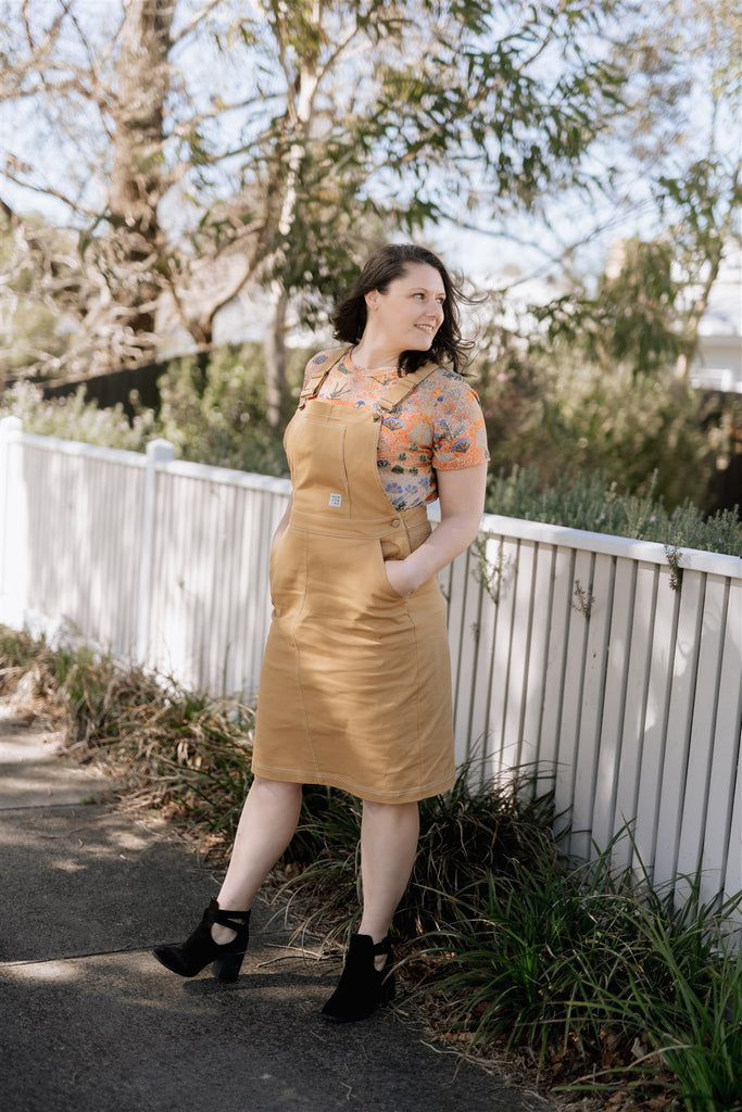 Bec K wears the Nancybird Overall Dress in Mustard and the Apollo Tee in Desert Flower print. She is smiling looking to her left while the wind blows her hair gracefully. The backdrop is eucalyptus leaves and a white and cream wooden fence.