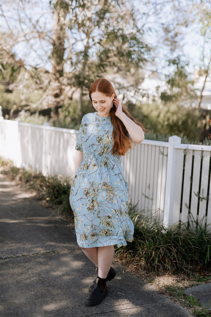 Bec S stands, smiling at the ground while tucking her long, red hair behind her ear. She wears the Nancybird Pepper T-Shirt Dress in Treetops and brown Blundstone Chelsea boots. The dress has a pale, sky blue background with a painting of gumtree branches, leaves and cockatoos printed on it.