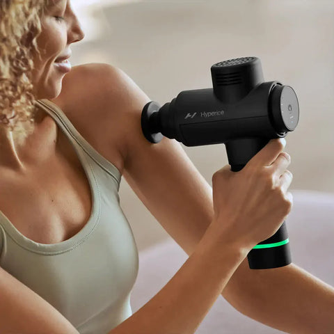 Una mujer usa la pistola de masaje portátil Hyperice HyperVolt 2 para aliviar el dolor en casa.