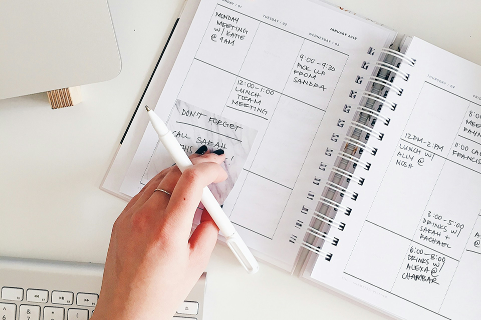 keyboard and daily planner with notes for each day, and a hand holding a pen in the foreground. 