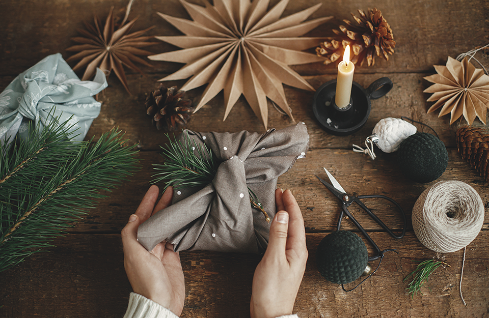 Hands holding stylish xmas gift wrapped in brown fabric on rustic wooden table with scissors, paper star, candle. 