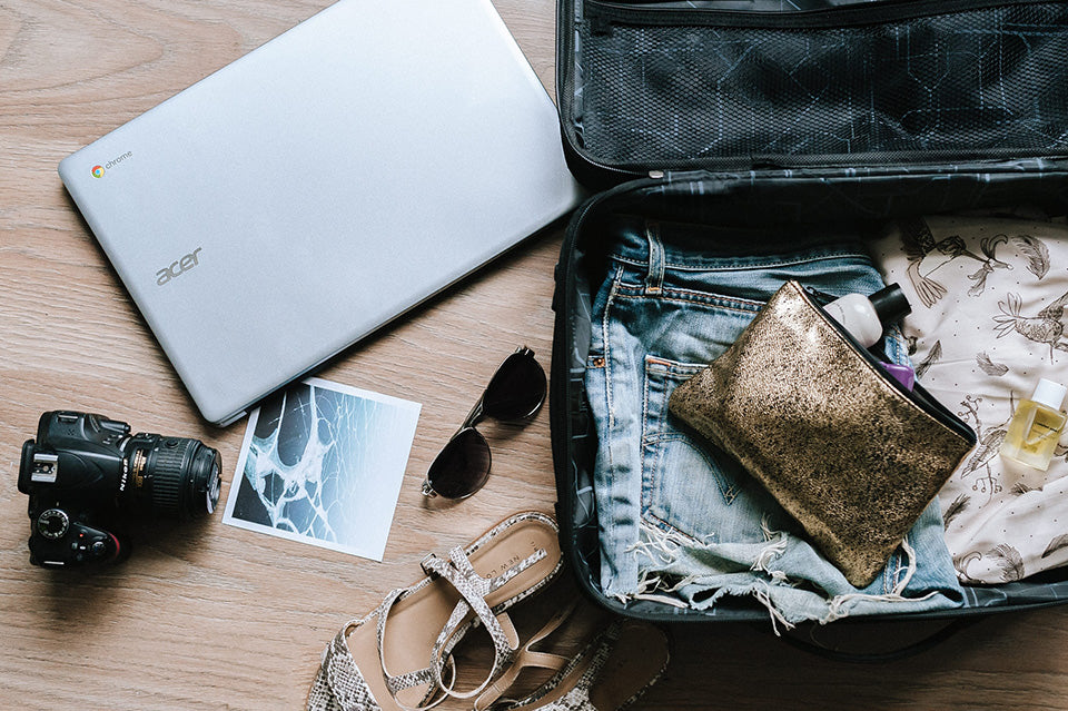 An open suitcase containing a pair of short jean shorts, a patterned shirt, a small bag with toiletries. Outside the suitcase are a Chromebook, a camera, a postcard, sunglasses, and sandals.