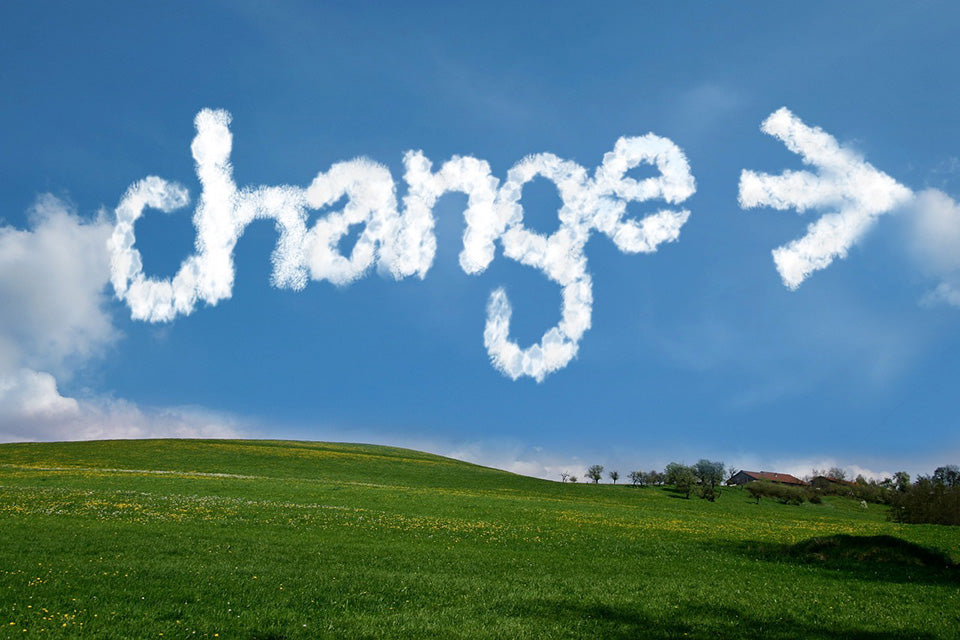 A green rolling pasture with a house on top of the hill. In the perfectly blue sky clouds are arranged to say 'change' and an arrow pointing right.