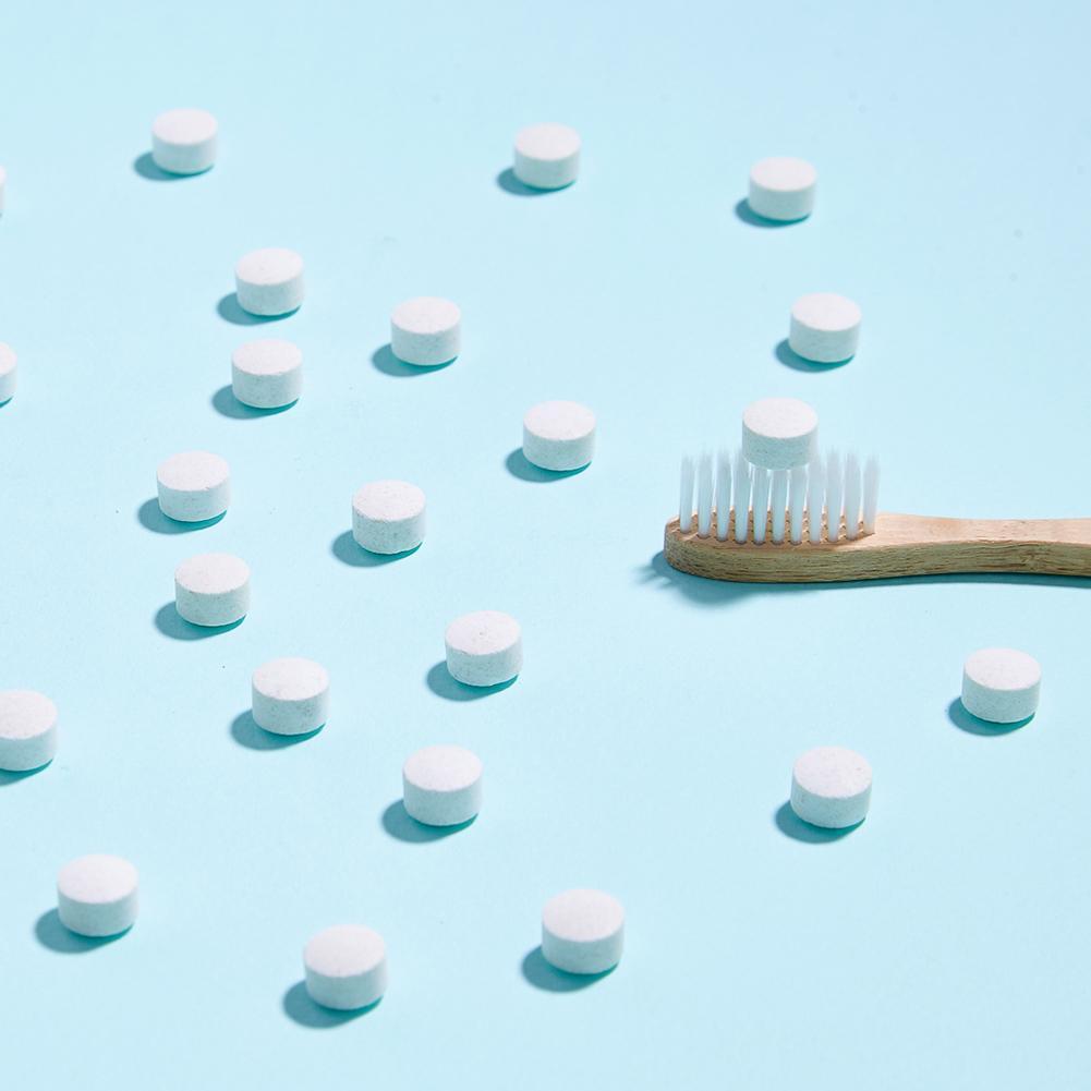 Blue background with tooth tablets sprinkled all around and one tablet on top of the head of a bamboo toothbrush