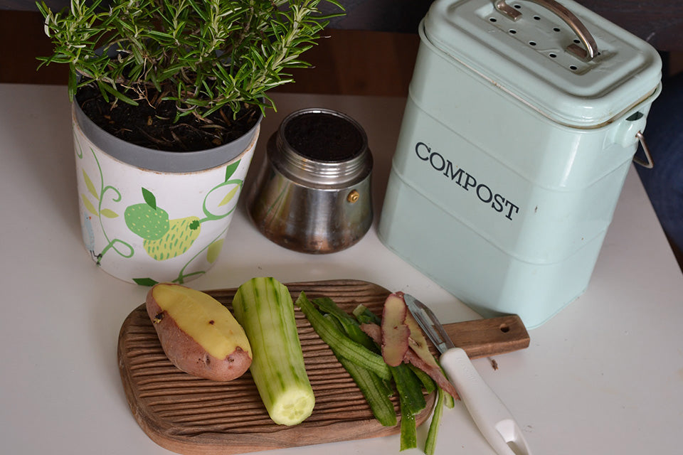 : A cutting board with a peeled potato and peeled zucchini sitting on top next to their peels. Behind the board is a tin countertop compost bin, the bottom of a bialetti, and a plant