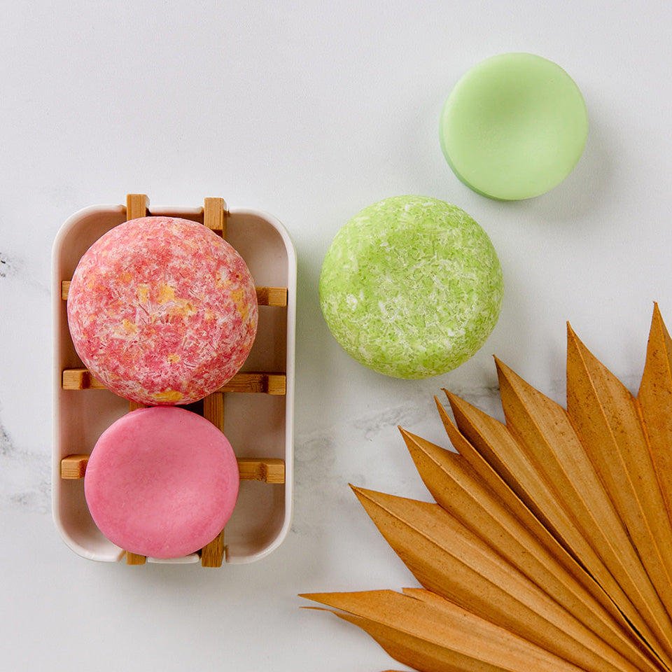 A pink shampoo and conditioner bar pair on a soap dish next to a green shampoo and conditioner bar pair