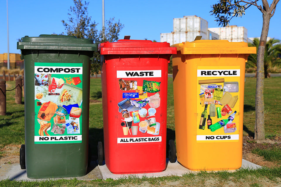 Three bins in a public space standing next to each other. From left to right they are labeled Compost - No Plastic; Waste - All Plastic Bags; Recycle - No Cups.