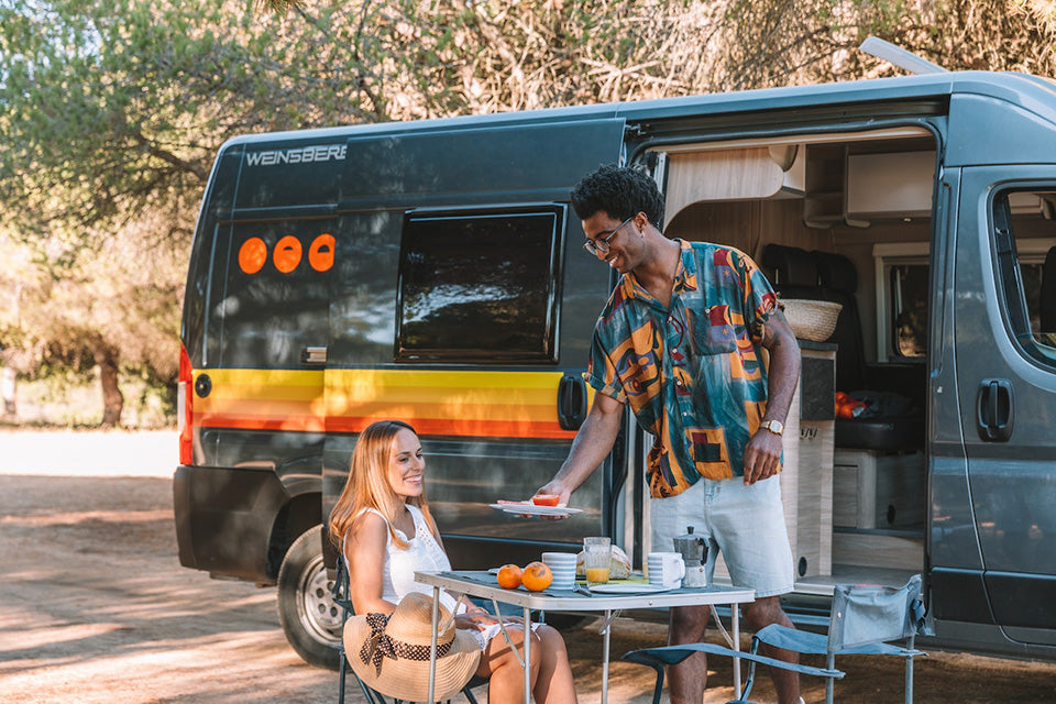 man in colorful polo shirt serving woman in white a plate of food outside of a camper van