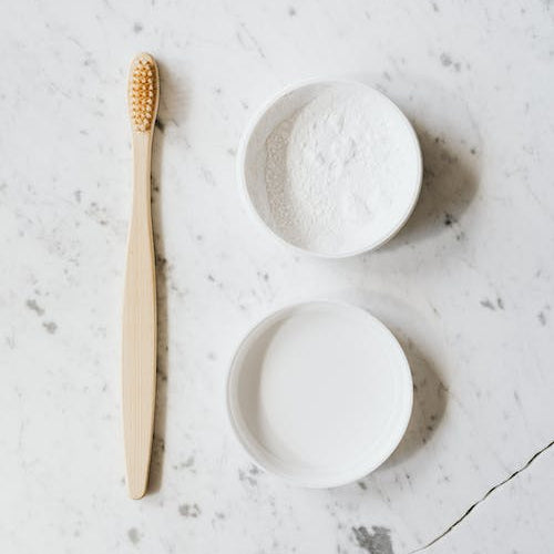 A bamboo toothbrush sits on a stone countertop next to an open white container holding tooth powder.