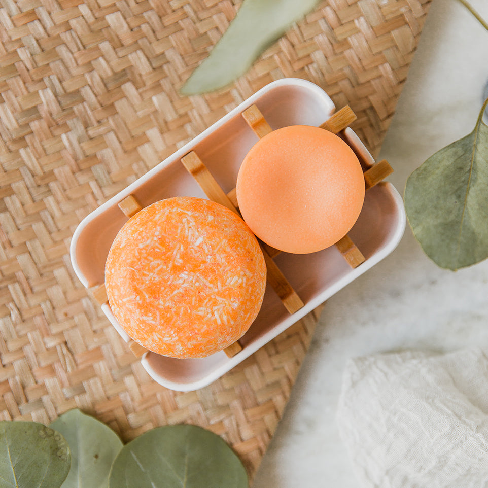 A set of orange sunshine shampoo and conditioner bars on a white soap dish with bamboo slats.