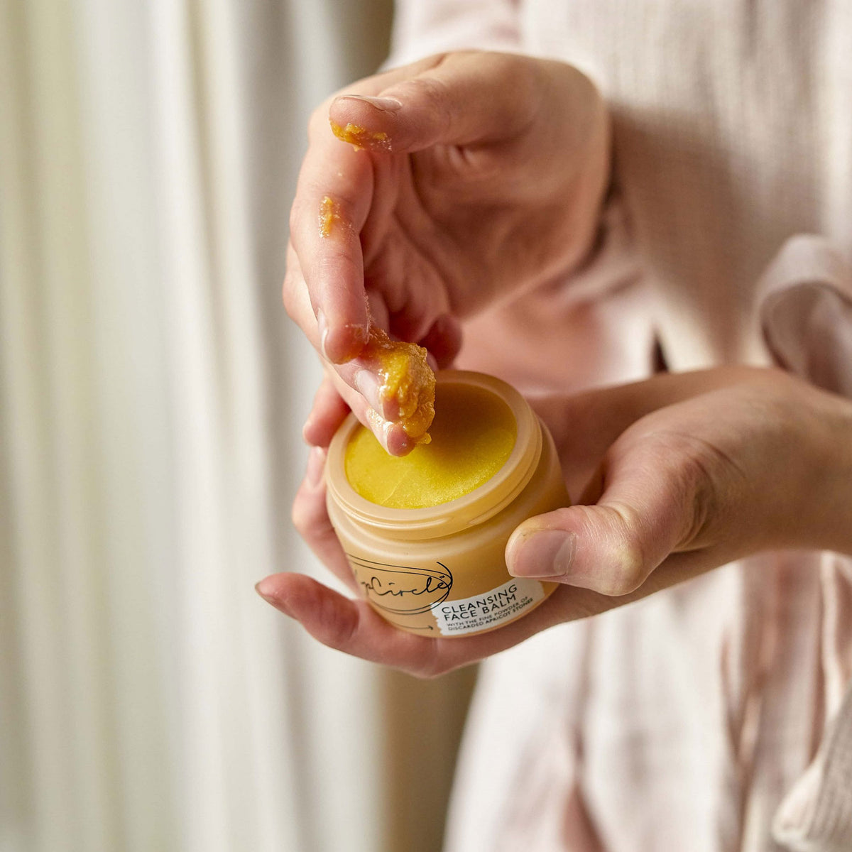 Person holding a jar of the yellow cleansing face balm in one hand, scooping out a quarter sized amount onto fingers with the other hand.
