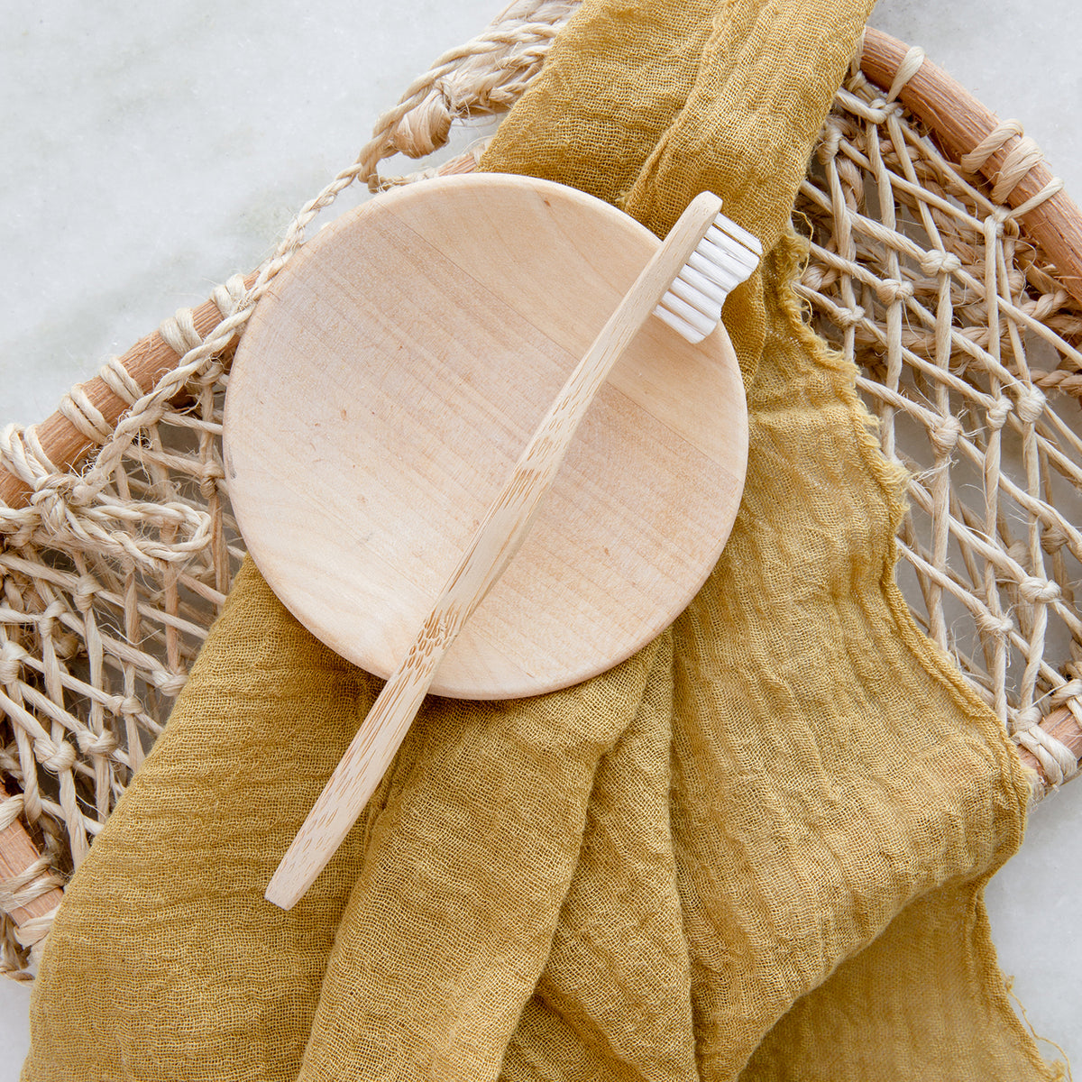 A bamboo toothbrush resting on a small circular bamboo dish and an arrangement of a mustard cloth and natural woven object