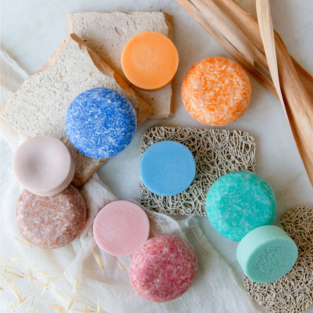 A collection of orange, blue, tan, pink, and turquoise shampoo and conditioner bars arranged on soap lifts, white muslin cloth, and small slabs of stone 