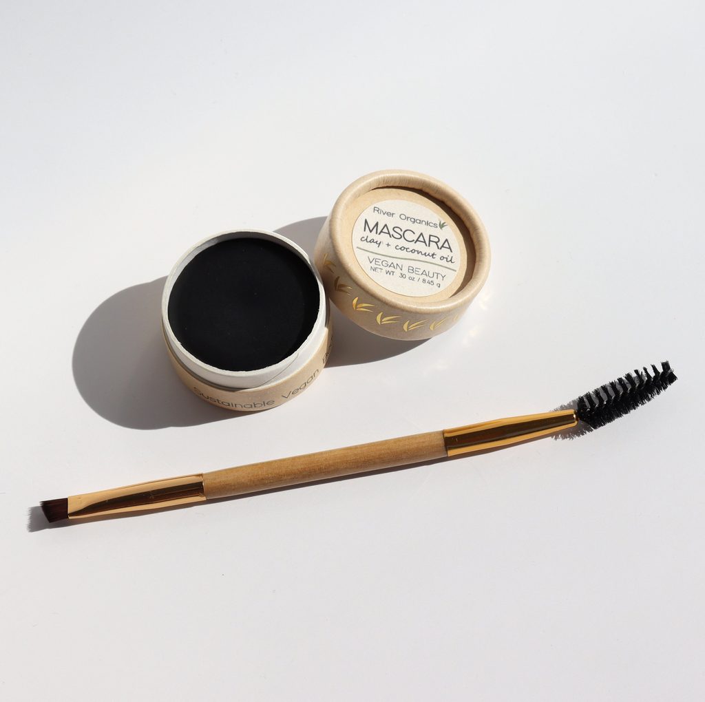 Small cardboard circle container containing solid black mascara. Life off to the side reads: River Organics Mascara. Mascara spoolie and eye shadow applicator combo brush sits in front