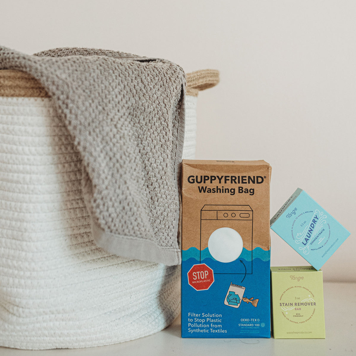 A boxed Guppyfriend Washing Bag, a boxed stain remover bag, and a boxed laundry concetrate next to a cloth laundry hamper with a gray towel hanging over the side