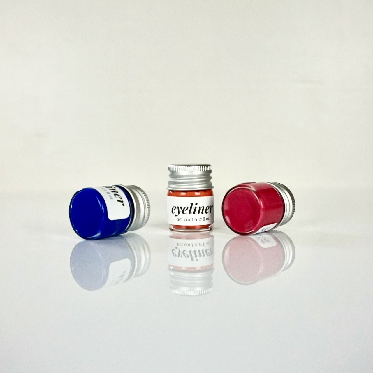 3 liquid eyeliners in small glass jars with tin lids in blue, orange, and red