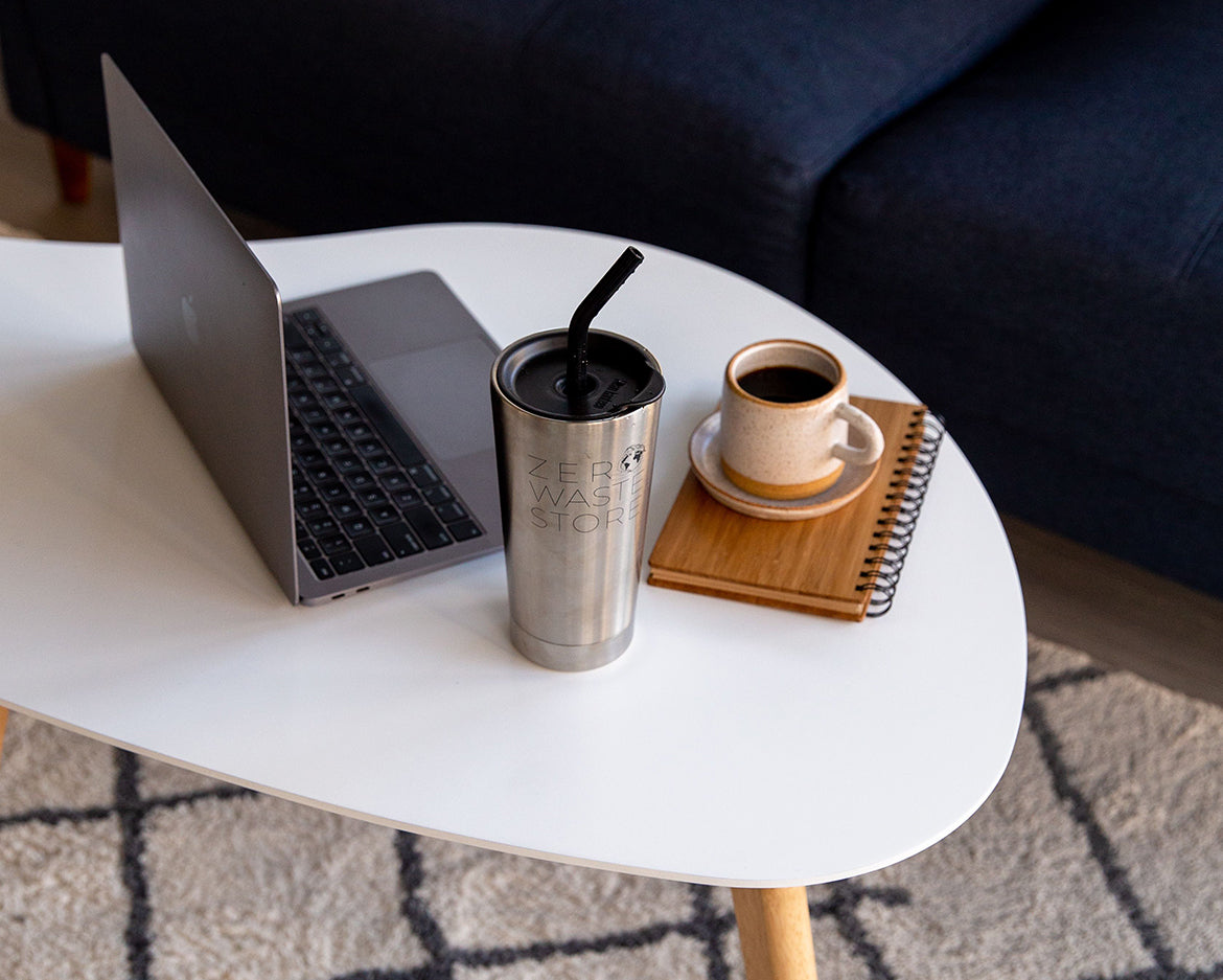 A laptop, a reusable stainless steel tumbler with a silicone straw cap, a small ceramic coffee cup and saucer, and a bamboo cover notebook on a white kidney-shaped coffee table