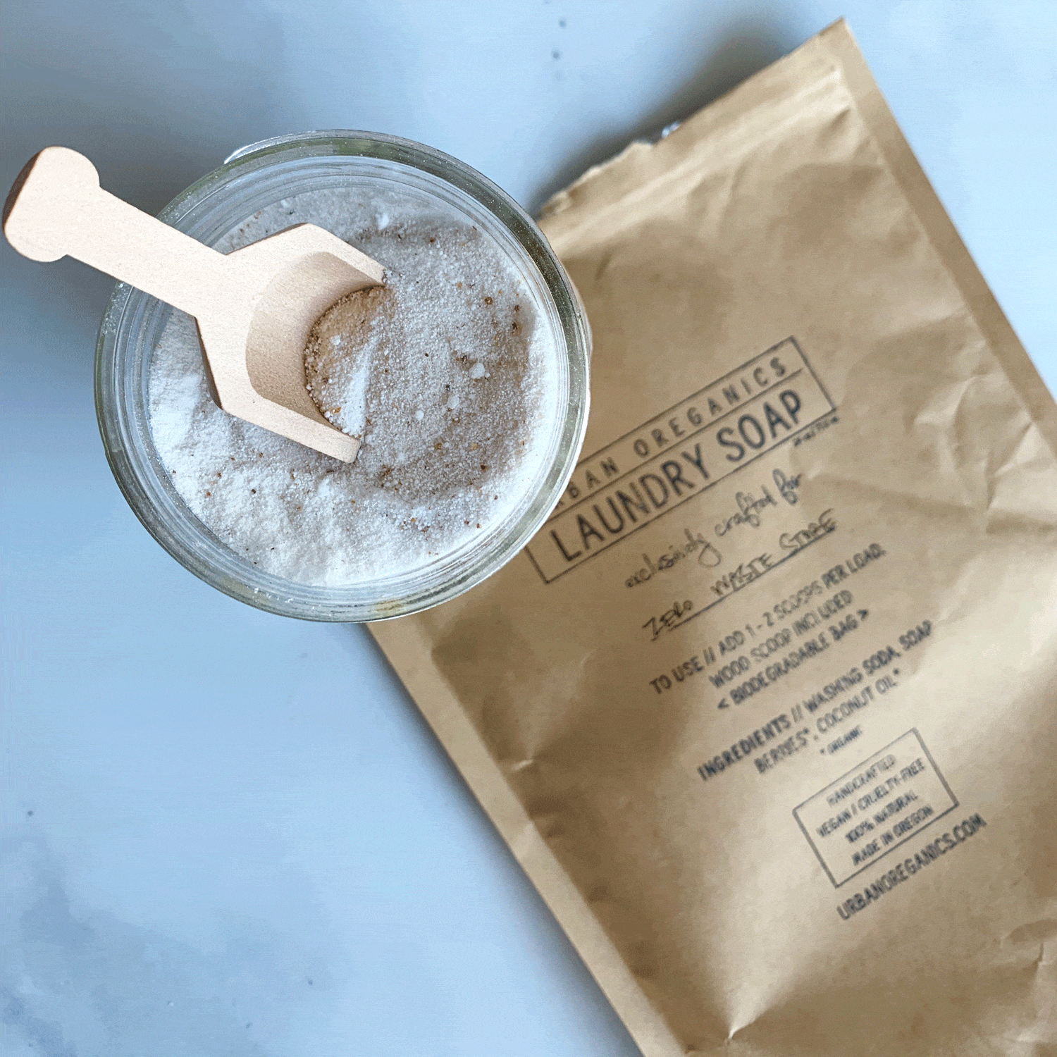 Glass bowl of laundry detergent powder with wooden scoop inside sitting on paper bag. Paper bag reads Urban Oreganics Laundry Soap.