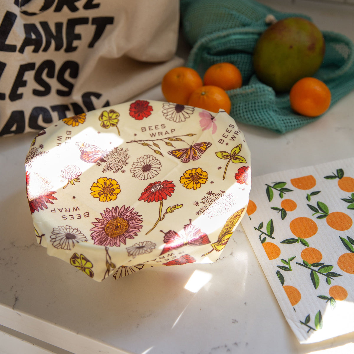 a bowl wrapped with flower print bees wrap, an orange print sponge cloth, and in the background a blue net bag of fruits