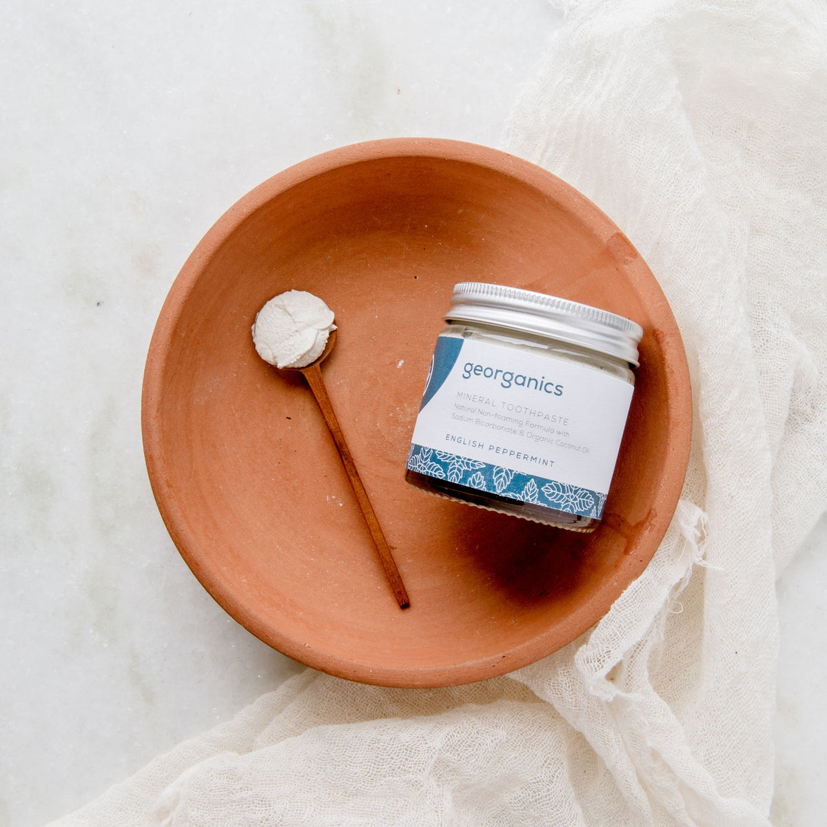 Georganics glass jar of toothpaste in a terra cotta bowl next to a bamboo spoon full of toothpaste