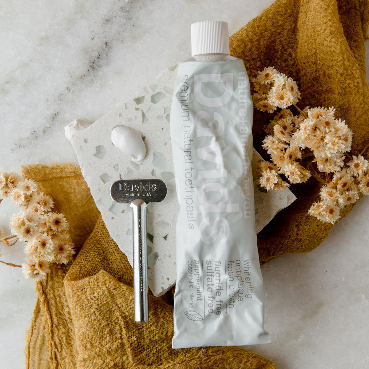 A metal tube of toothpaste, metal tube key, and small drop of white toothpaste balancing on a small slab of terrazzo tile, surrounded by small dried flowers and a mustard linen