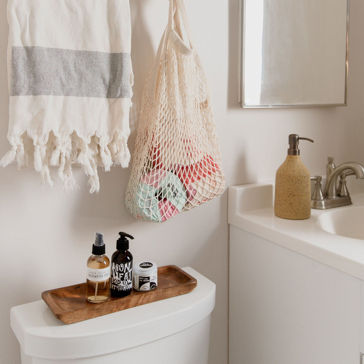A white bathroom interior with a tasseled towel and netted bag holding toilet paper hanging on the wall, a wooden tray of skin care products placed on top of the toilet, and to the right a ceramic soap dispenser next to a sink