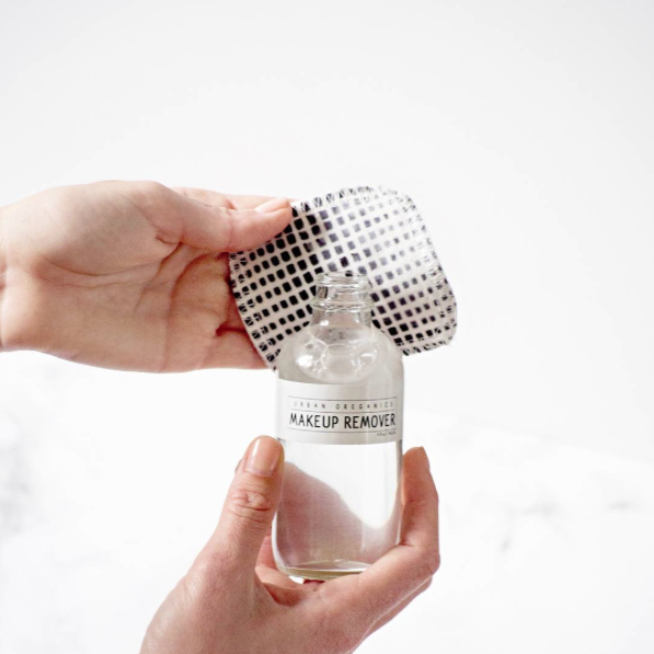 Hands holding a clear glass jar of open makeup remover upright with a reusable cotton square behind it.