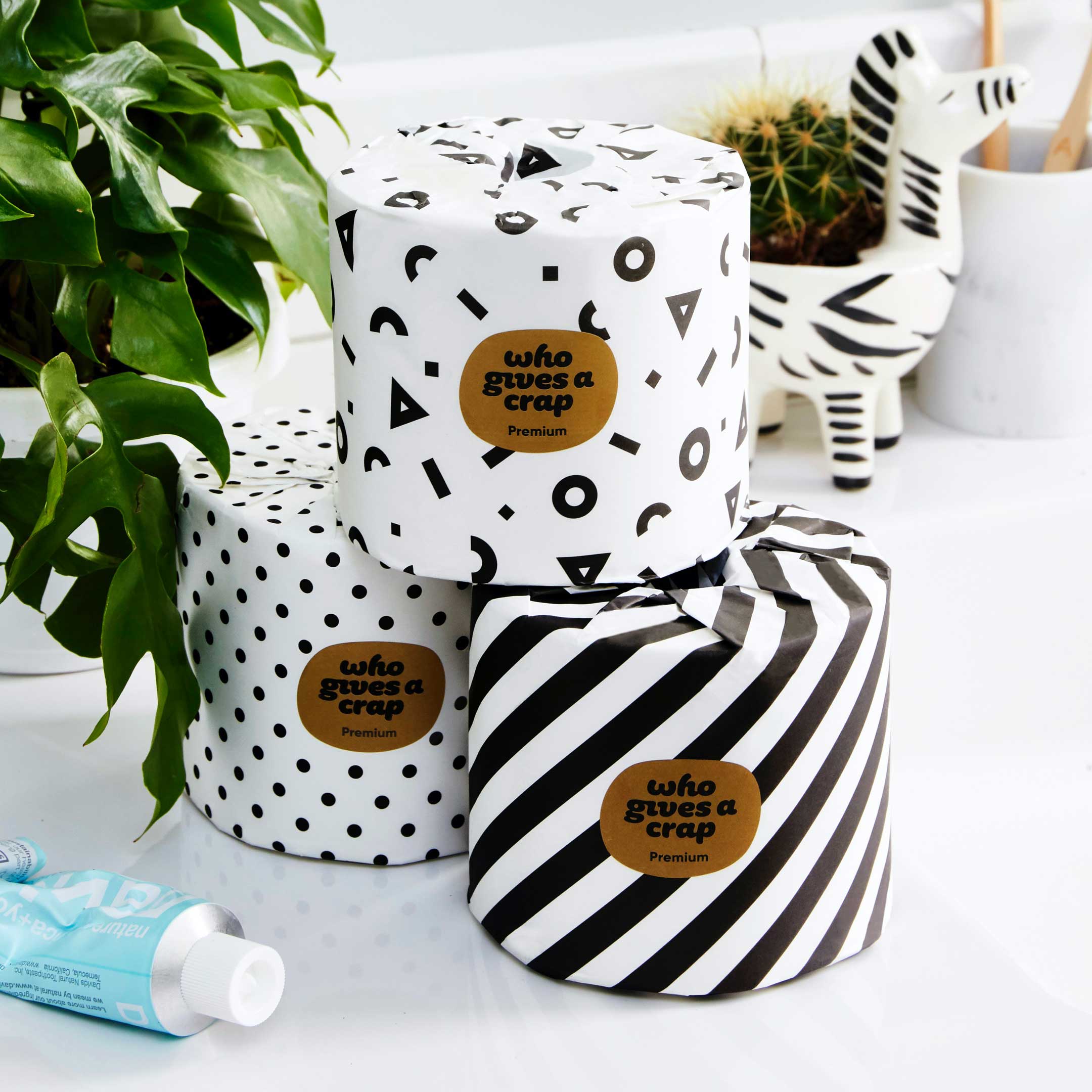 3 individually wrapped toilet paper rolls sitting on vanity with Who Gives A Crap Label. Patterns are black and white and include a polka dot and striped design