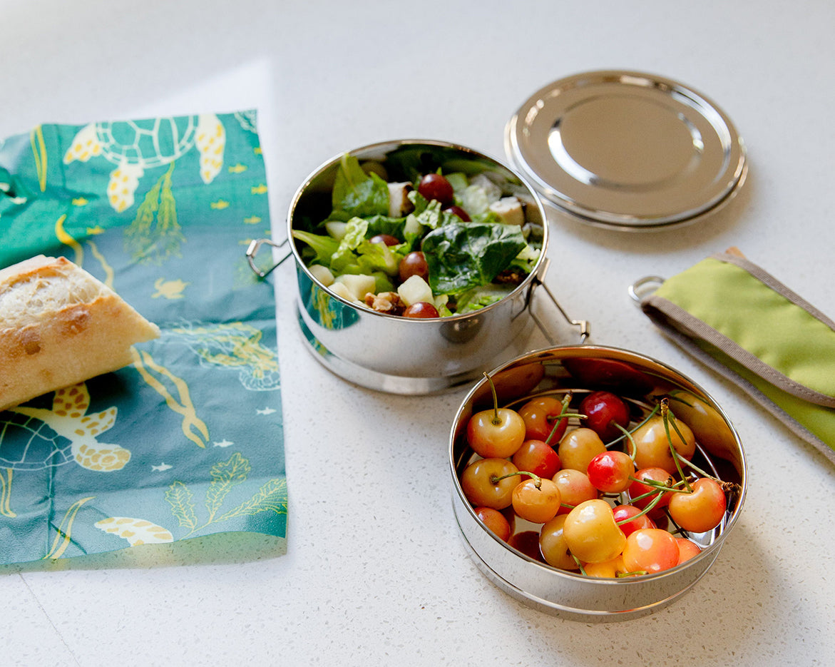 Two round stainless steel lunch boxes filled with salad and Rainier cherries. To the left is part of a baguette on a wax wrap, and on the right is a green utensil sleeve