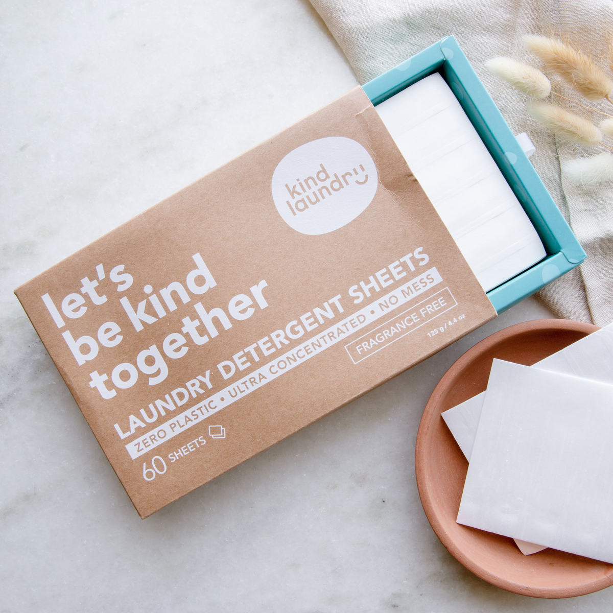 Cardboard box with blue insert pulled partially out and white sheets inside. 2 White sheets are outside box on clay plate. Box reads: let's be kind together laundry detergent sheets. Zero plastic ultra concentrated no mess.