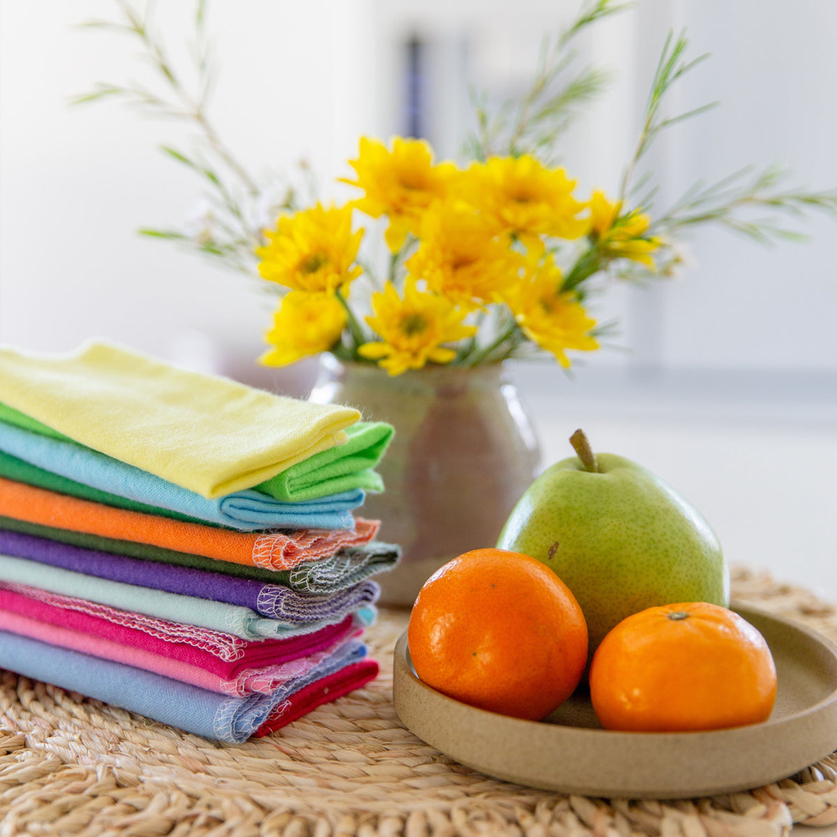 Stacked rainbow cloth towels and a ceramic plate of fruits on a raffia woven mat, a vase of yellow flowers in the background