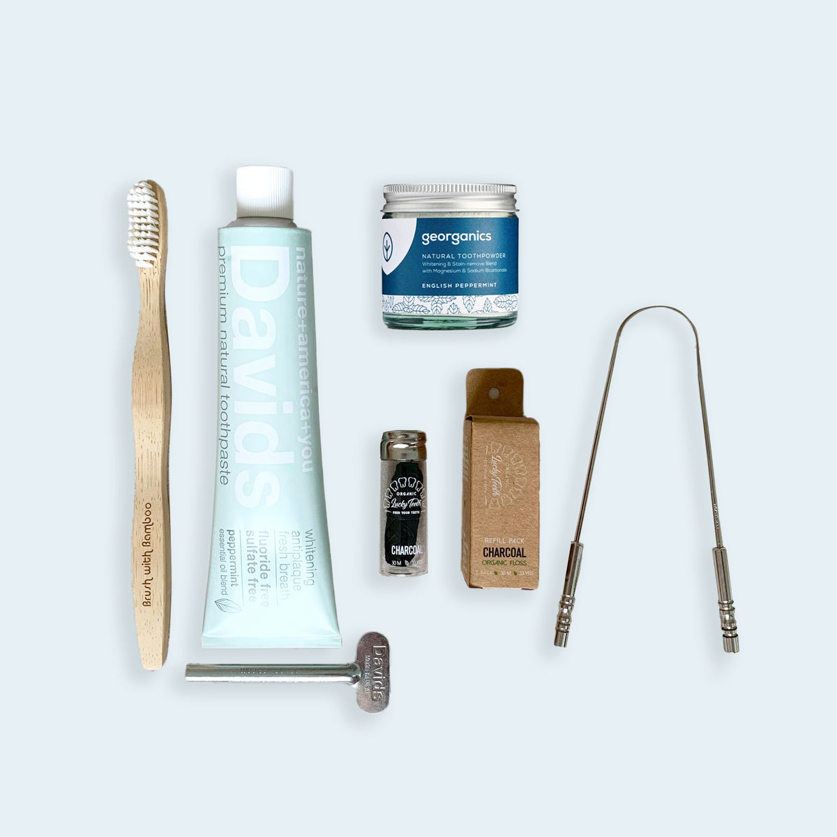 From left to right bamboo toothbrush, david's toothpaste tube with tube key underneath it, georganics tooth powder jar, bamboo charcoal floss in glass reusable jar, bamboo charcoal floss refill in cardboard box, and stainless steel tongue scraper