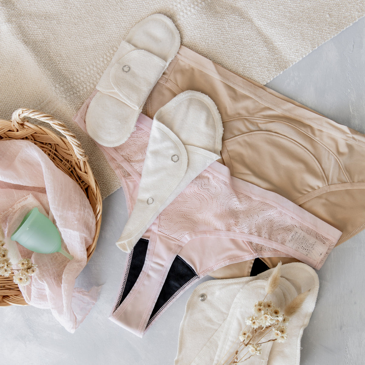 A selection of menstrual cloth pads and pantyliners, a mint-colored menstrual cup, and pink and nude menstrual underwear on a white surface.