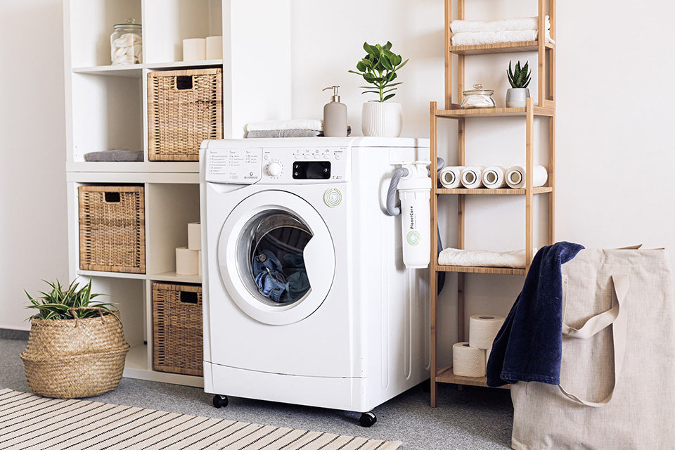 A washing machine is washing a load of clothes with a bag of laundry sitting next to it.