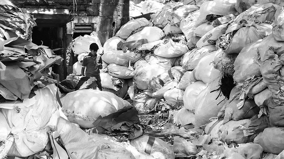 small boy surrounded by bags of trash on both sides of him 2x as tall as he is.