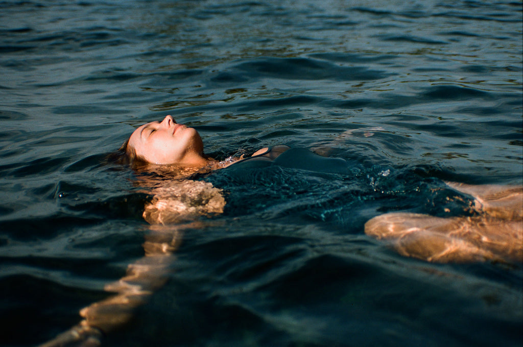 Sierra floating on her back in water, wearing a one piece surf swimsuit