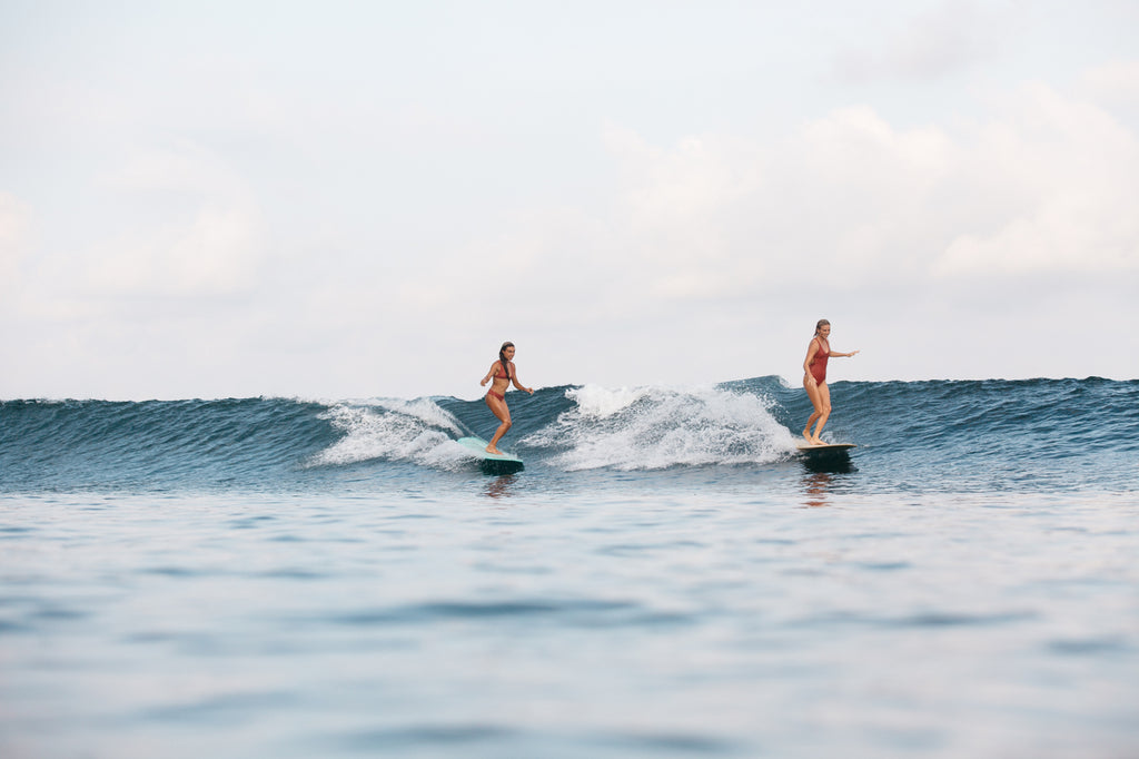 Two girls surfing a party wave. One wearing a surf bikini and the other wearing a one piece surf swimsuit in burnt sienna