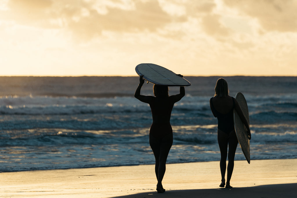 Two women walking to the water carrying their surfboards at sunset