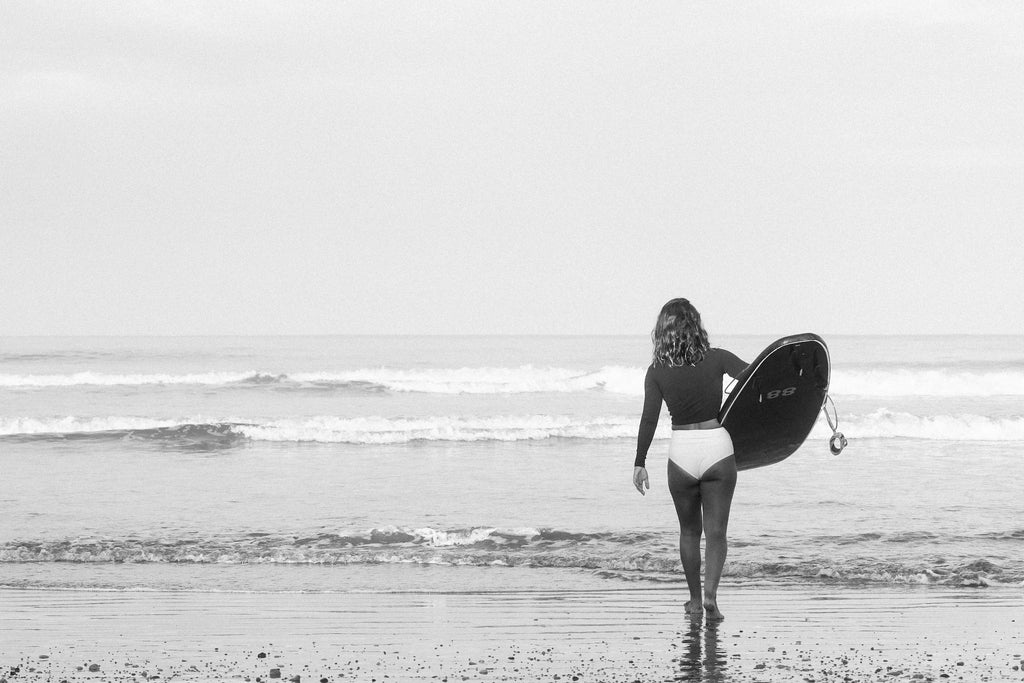 Erika walking to the water with surfboard under her arm wearing rash guard and lucky high waisted bikini surf bottoms