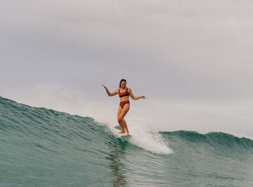 Woman doing a cheater five on her surfboard wearing the mason surf bikini top and kennedy surf bikini bottom in rose