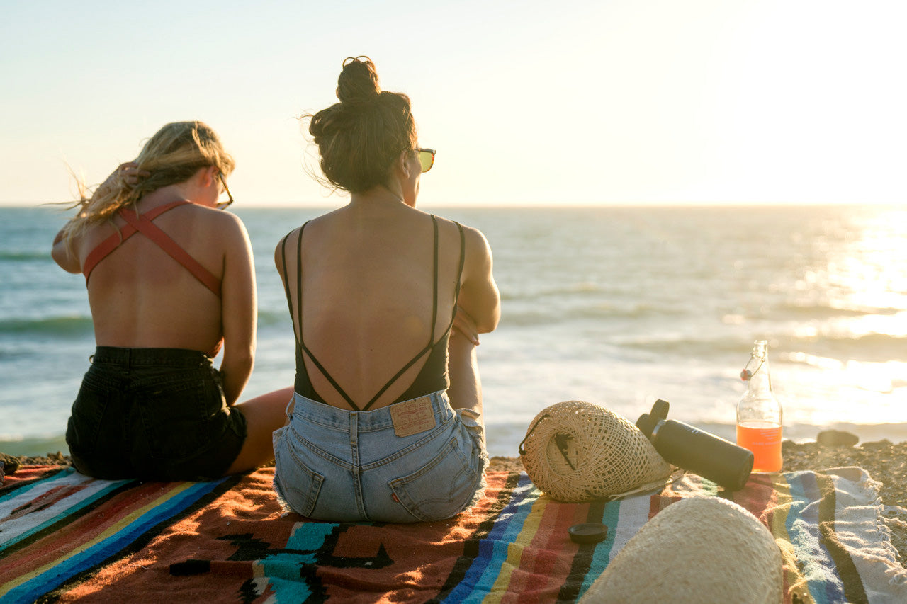 Two women sitting on the edge of a cliff watching the sunset having a picnic wearing jean shorts and one piece surf swimsuits