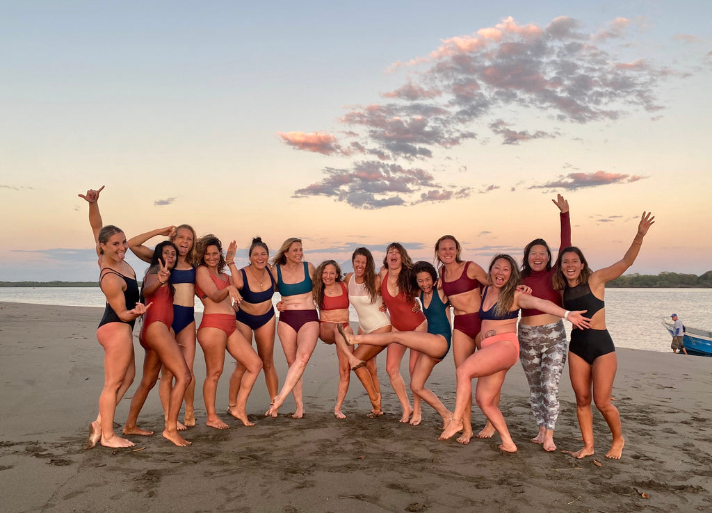 group of 15 women wearing surf bikinis and one piece swimsuits standing on a beach with arms around each other some with their hands in the air 