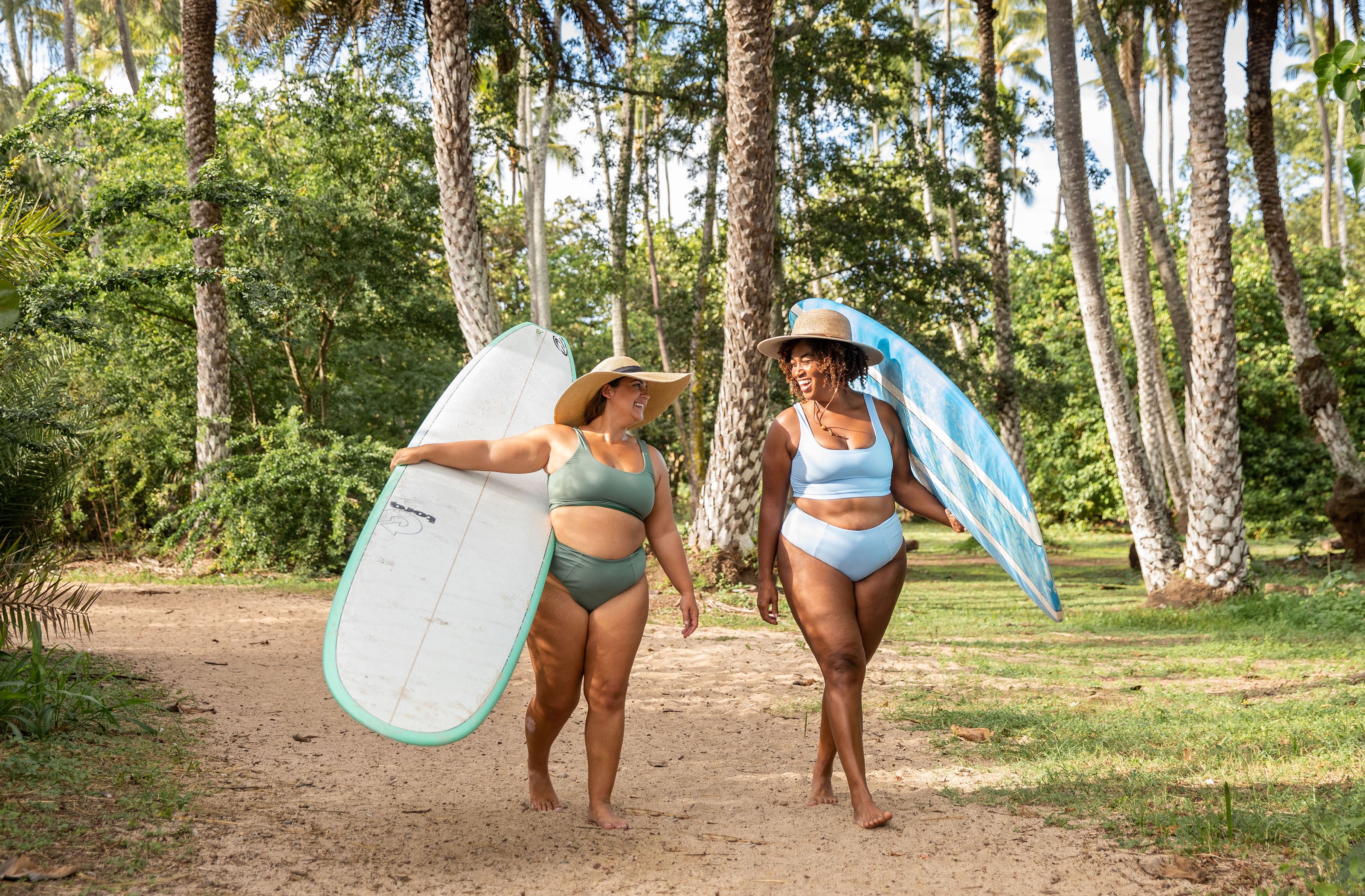 Two women walking down path carrying long board surf boards