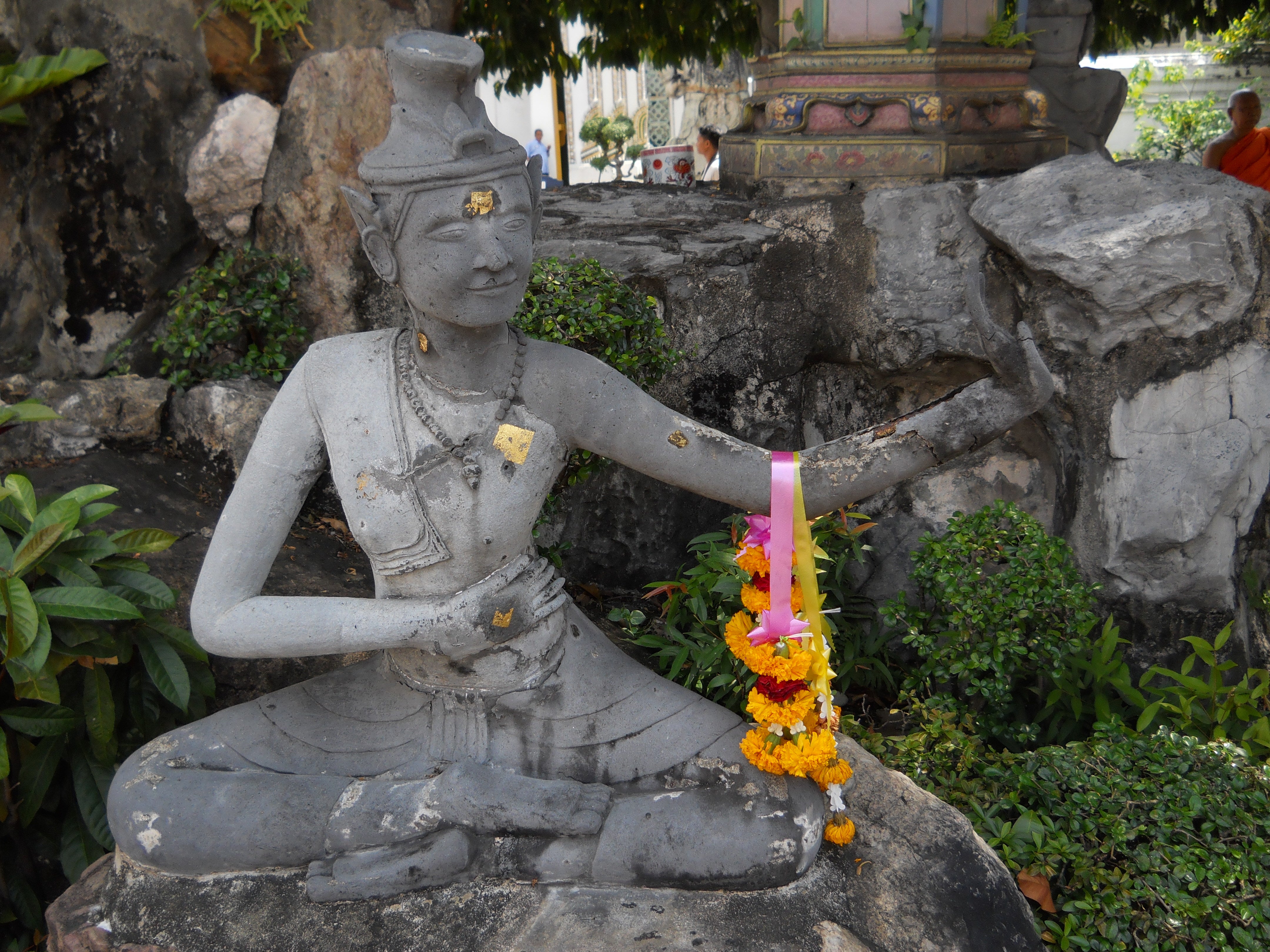 Concrete statue of a Thai person performing a Thai yoga pose, seated, with arm outstretched, and a flower garland draped over the arm.