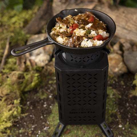 A tall, black outdoor stove on dirt and grass. A pan with food inside is on top, cooking.
