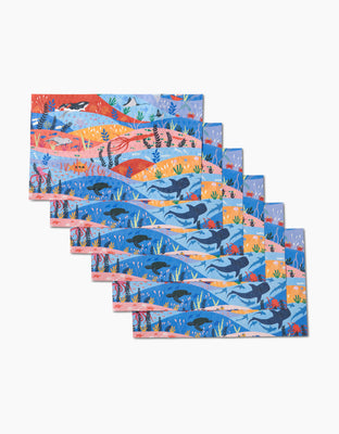 Oceans by Ceyda Alasar features seven illustrated placemats overlapping in a fan shape, each showcasing vibrant ocean scenes with whales, fish, and sea plants on white, made from eco-friendly post-consumer recycled materials.