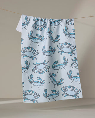 The Mindfulnice Coastal Crew tea towel, featuring a quick-drying white fabric with a repeating blue crab pattern, hangs on a line against a neutral background.